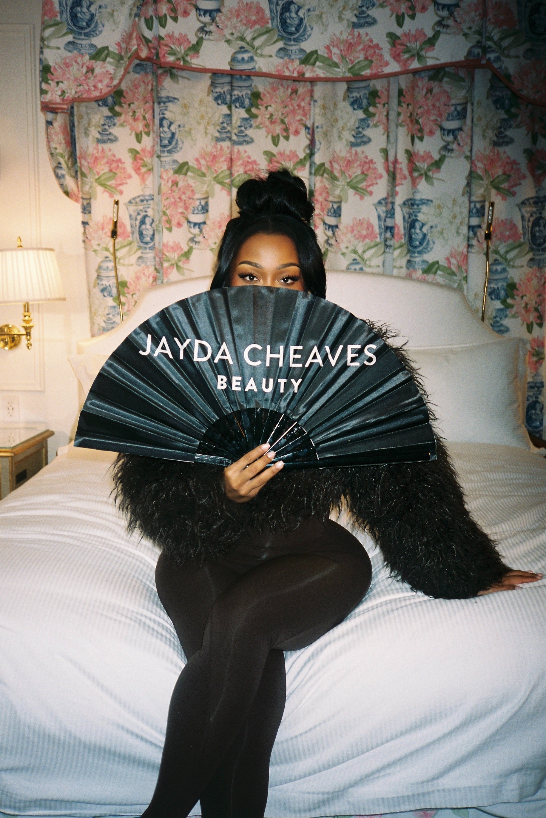 Jayda Cheaves holding a branded fan on a bed with floral curtains in the background
