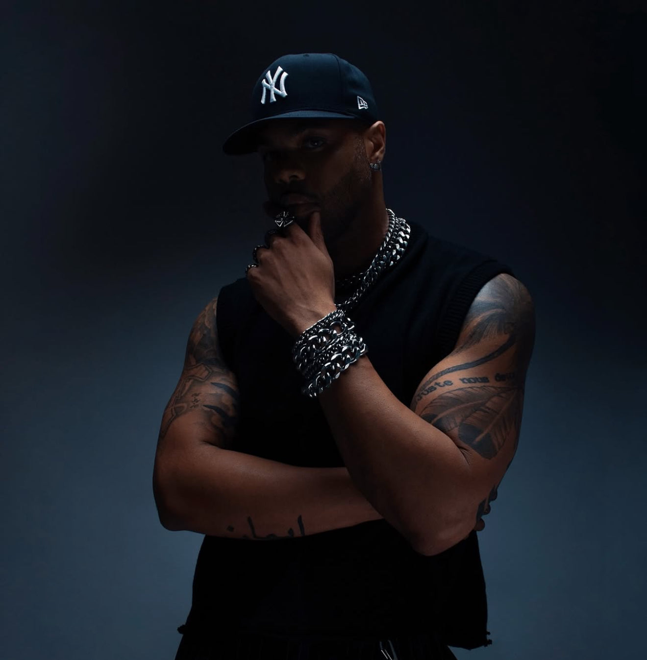 AX Rated wearing a black sleeveless shirt, dark cap, and multiple necklaces with tattoos on arms against a dark background
